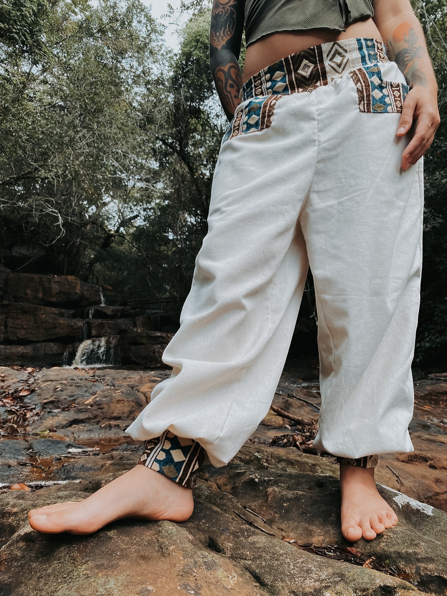 Goa Nomads cream linen pants with embroidered waistband and ankle cuffs featuring geometric patterns in brown and blue.